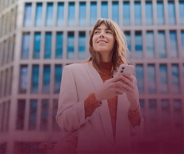 A smiling woman holding a mobile phone in front of a corporate building.